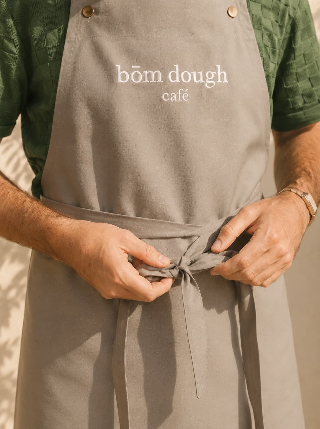 A bōm dough team member tying on a grey bōm dough café branded apron — one of the bakers and baristas in Cambridge.