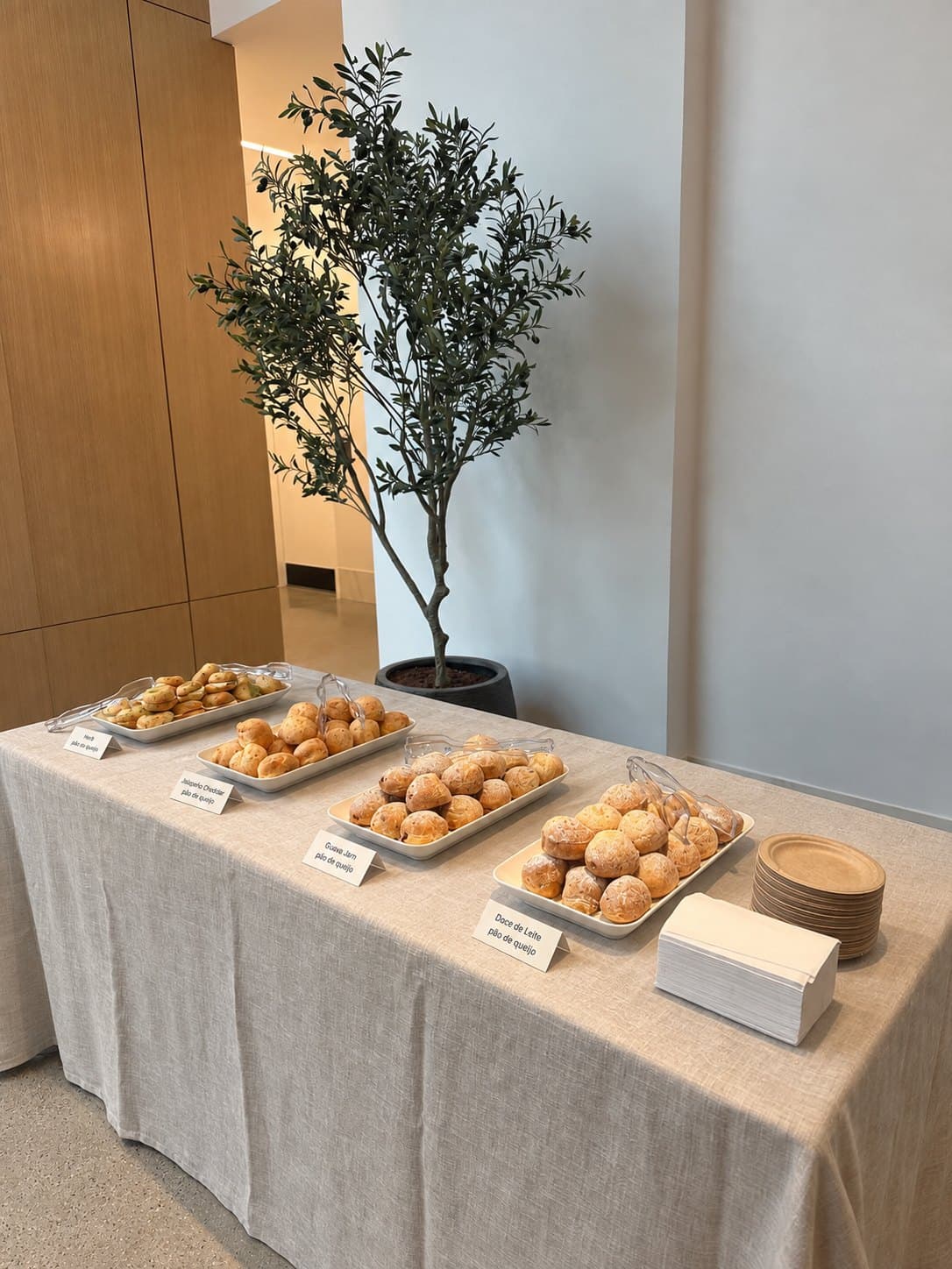 A bōm dough catering table — four white trays of pão de queijo (herb, jalapeño cheddar, guava jam, doce de leite) with tongs, labels, plates and napkins, on a linen-covered table in an office lobby.