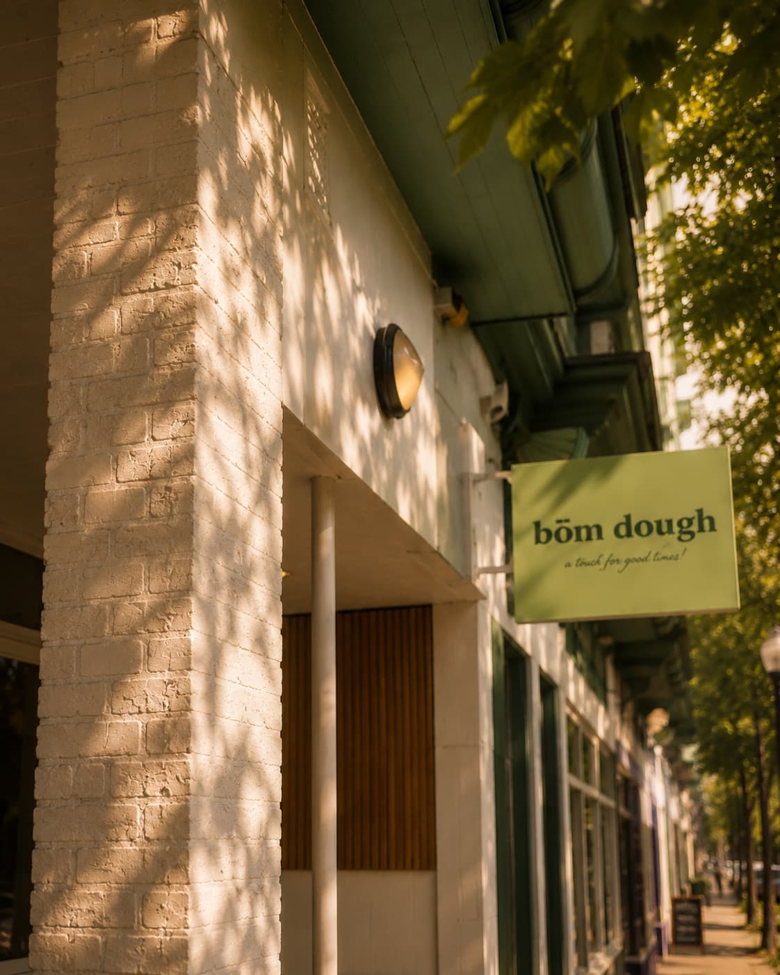 The bōm dough Inman Square storefront in Cambridge — a white-painted brick corner, the green bōm dough hanging sign, and dappled tree-light at golden hour.