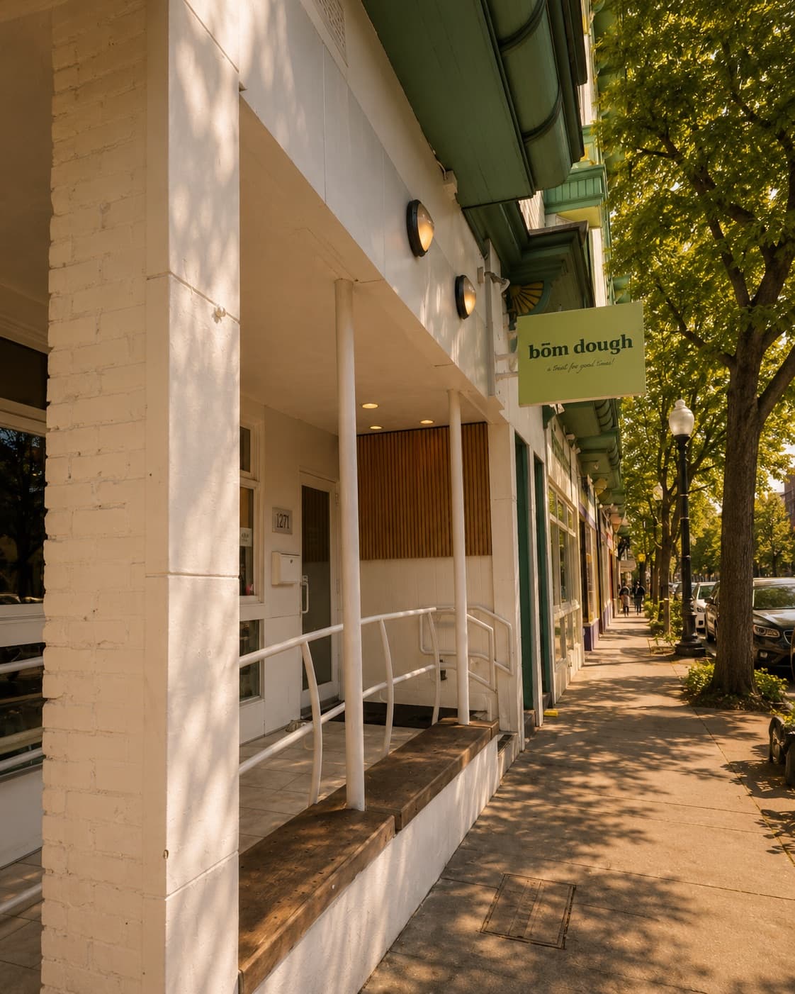 The bōm dough Inman Square café storefront at 1271 Cambridge St — a green-trimmed building with white columns, an accessibility ramp, and the green bōm dough · pão de queijo hanging sign on a tree-lined Cambridge sidewalk in golden light.