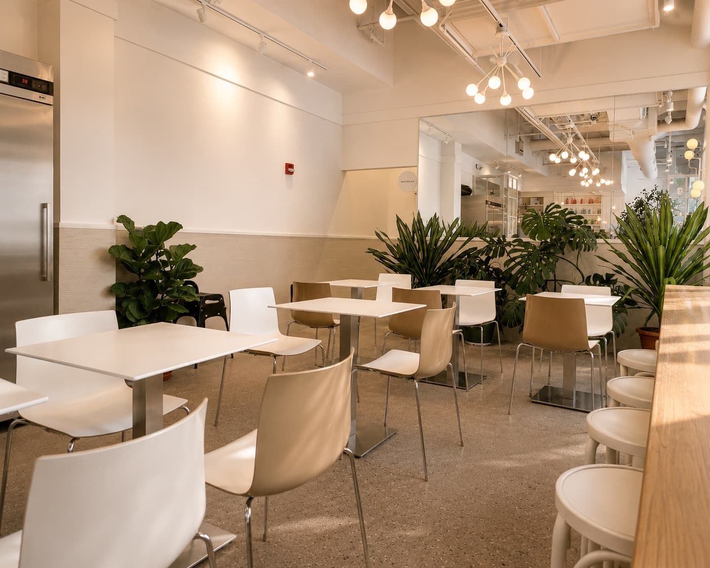 Bright interior seating at bōm dough Lechmere — white café tables and chairs, leafy plants, terrazzo floor, and pendant lighting.