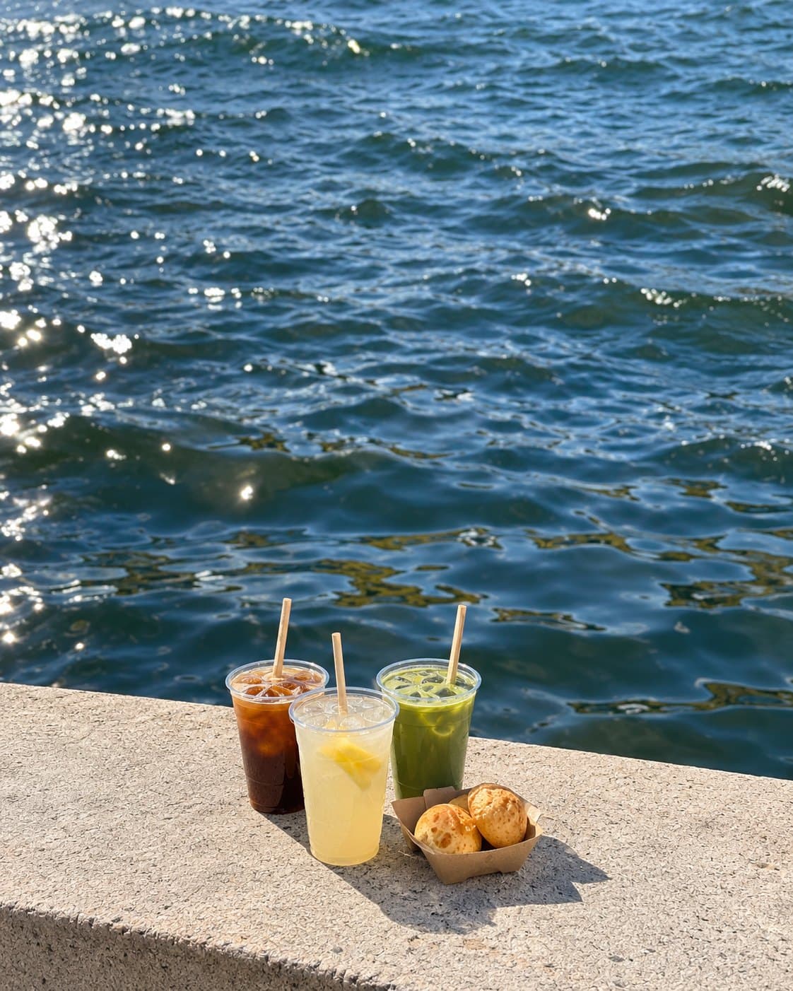 bōm dough iced coffee, matcha and fresh lemonade with pão de queijo — Lechmere, Cambridge Iced coffee, fresh lemonade and iced matcha beside a paper tray of pão de queijo on the granite edge of the Lechmere canal, Cambridge.